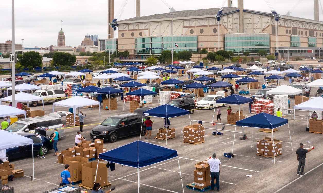 A photograph of a parking lot full of event tents with boxes of food under them -- volunteers serve up food to cars waiting in line.