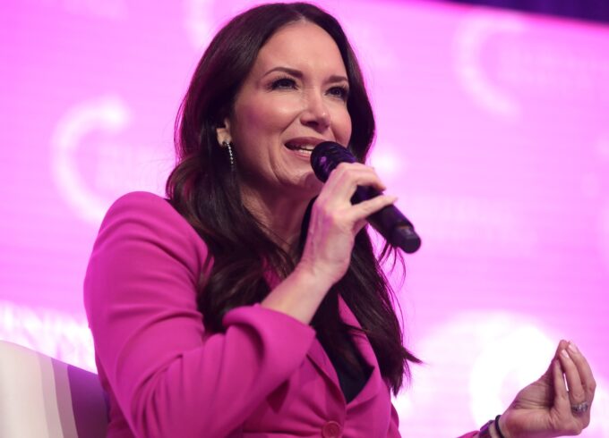 A photograph of Brooke Rollins wearing dark pink suit. She is seated in front of a pink background during a panel discussion.