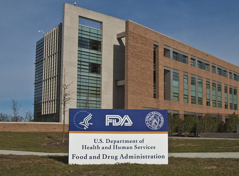 Outside shot of the U.S. Department of Health and Human Services Federal Drug Administration's Headquarters in Maryland. A sign out front says the name of the agency with its seal, the sign is white with blue text, yellow stripe and agency seal in white on a blue background. The background is a modern building made of concrete and red brick with a blue sky behind it.
