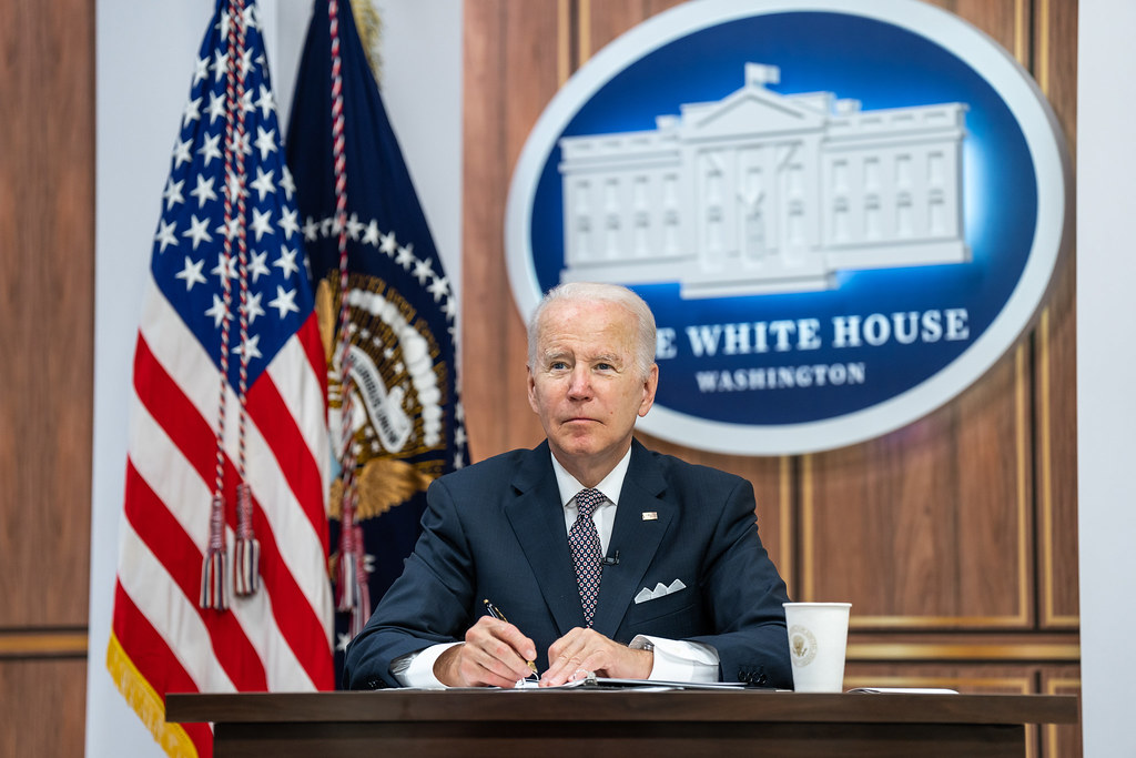 A photo of President Biden at a desk with a White House logo behind him.
