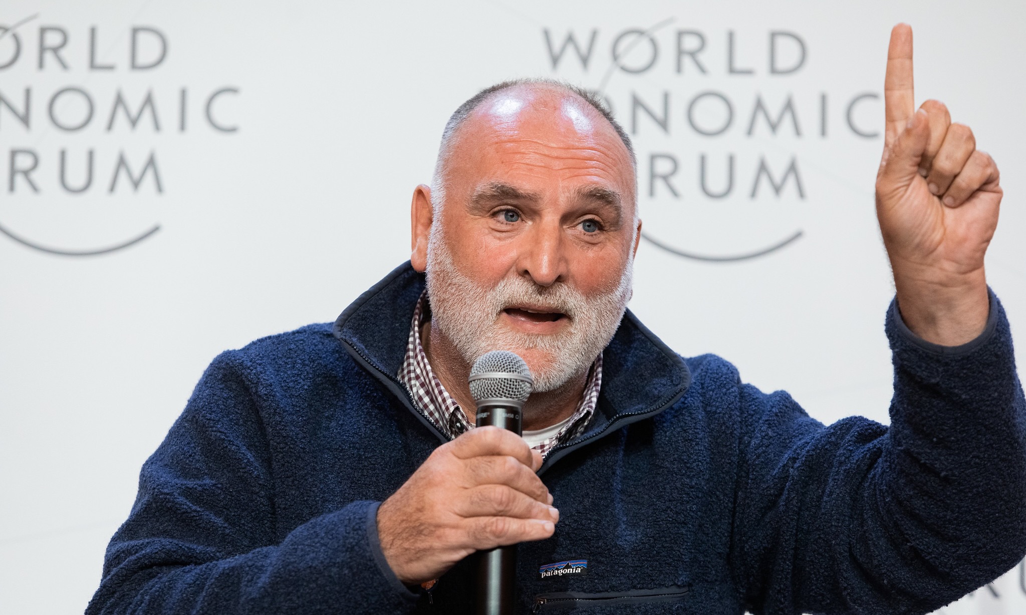 A photograph of humanitarian and chef José Andrés speaking at the World Economic Forum. He is wearing a navy blue patagonia fleece, donning a beard and holding a microphone and pointing a finger in the air.