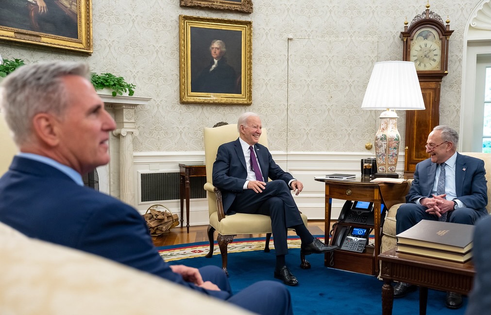 Photograph of Kevin McCarthy, Joe Biden and Chuck Schumer in the oval office. Biden is seated in the middle, McCarthy is out of focus in the foreground.