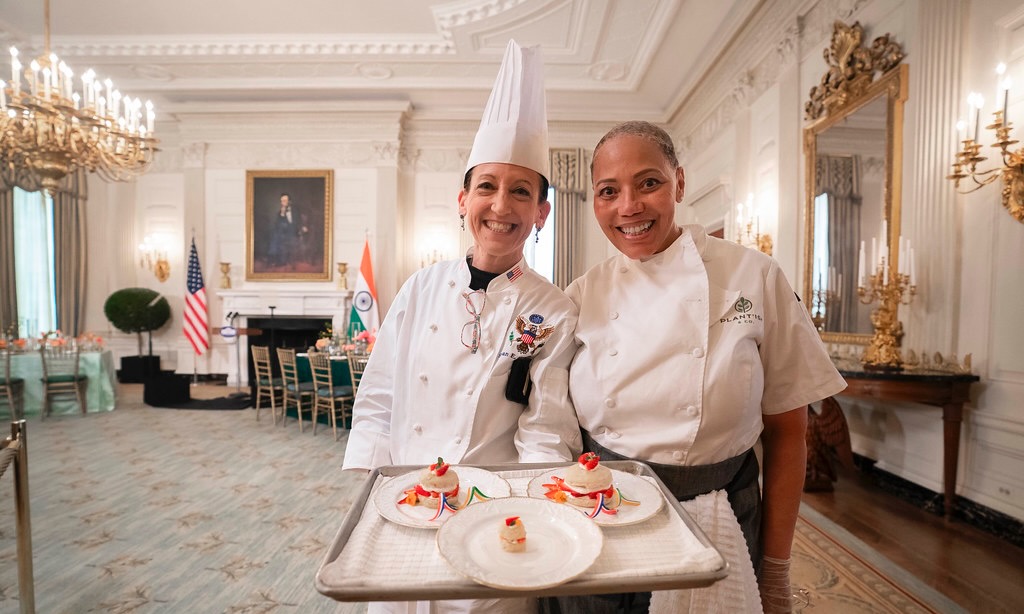 Two women White House chefs stand together posing for a photo inside a White House dining room. They are holding a tray with desserts.