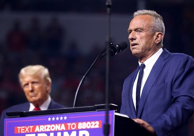 A photograph of Robert F. Kennedy Jr speaking at a blue podium with former President Donald Trump in the background.