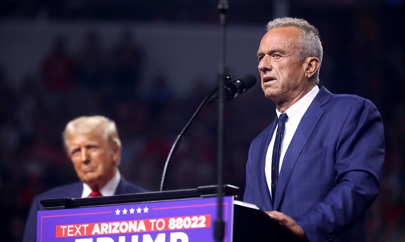 A photograph of Robert F. Kennedy Jr speaking at a blue podium with former President Donald Trump in the background.