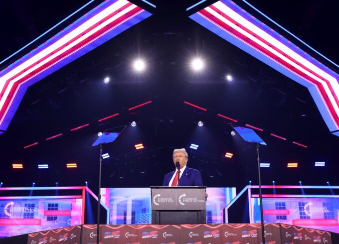 A photo of Donald Trump on a red white and blue stage in front of a podium with lights over head.