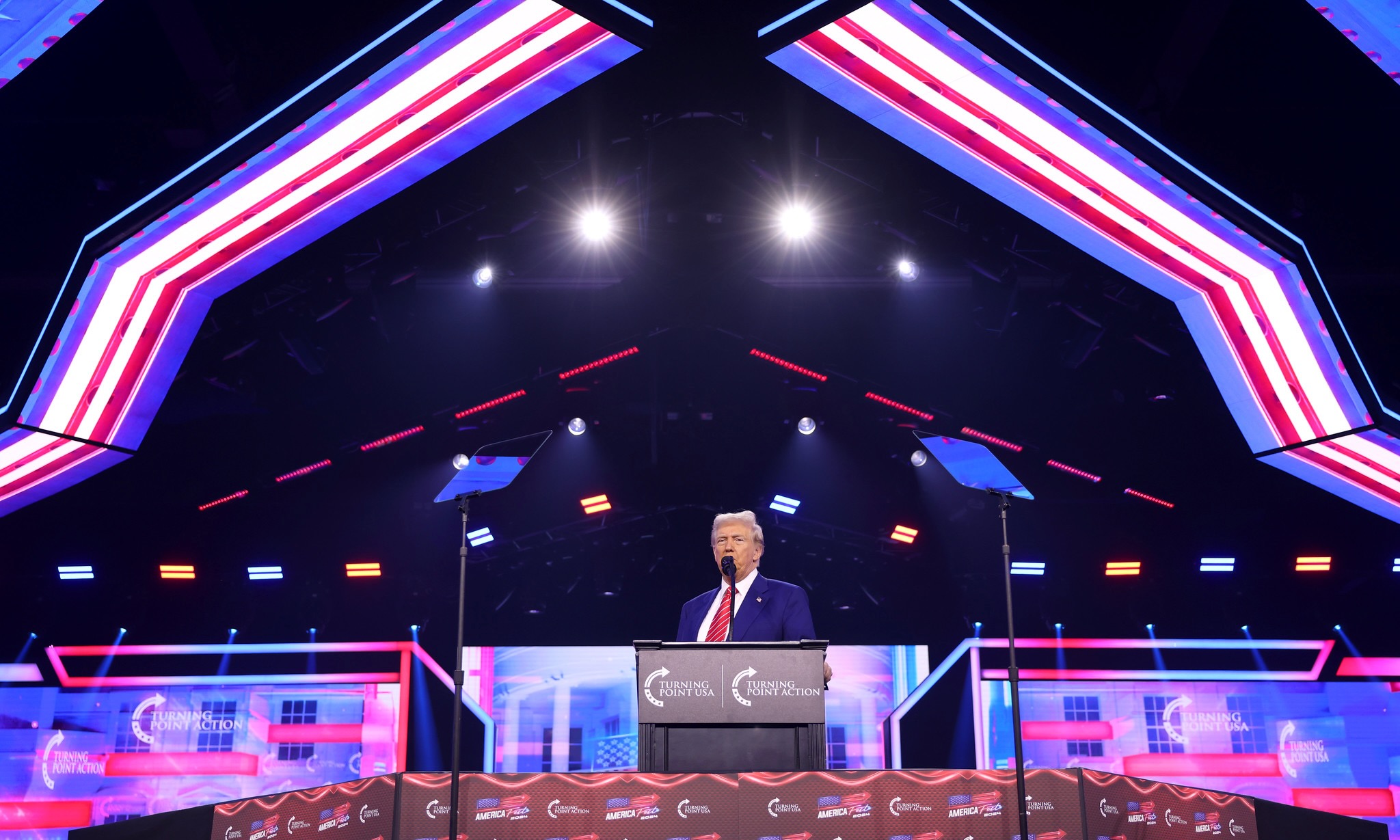 A photo of Donald Trump on a red white and blue stage in front of a podium with lights over head.