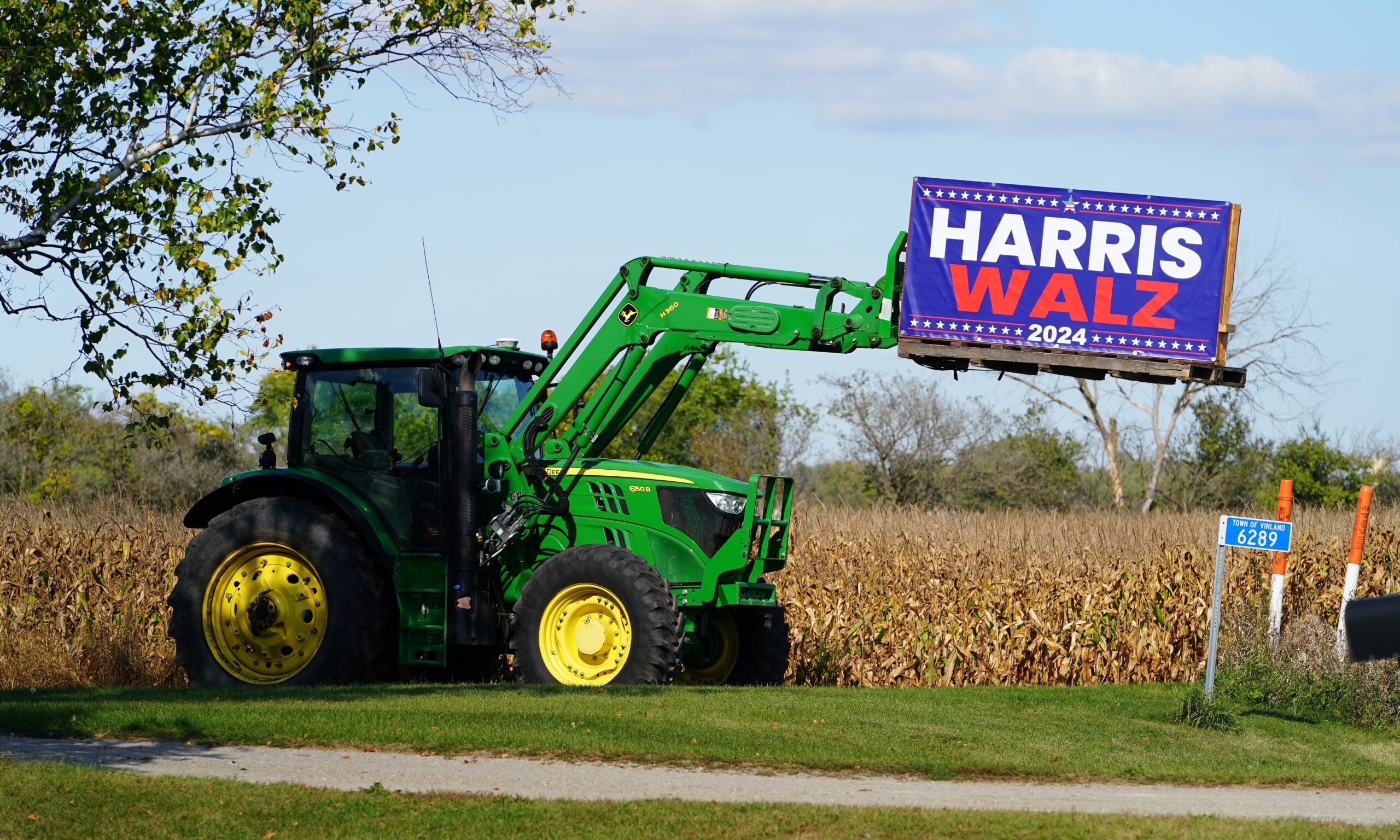 John Deere tractor holds a sign up supporting Kamala Harris and Tim Walz. The background is a farm with corn fields in view.