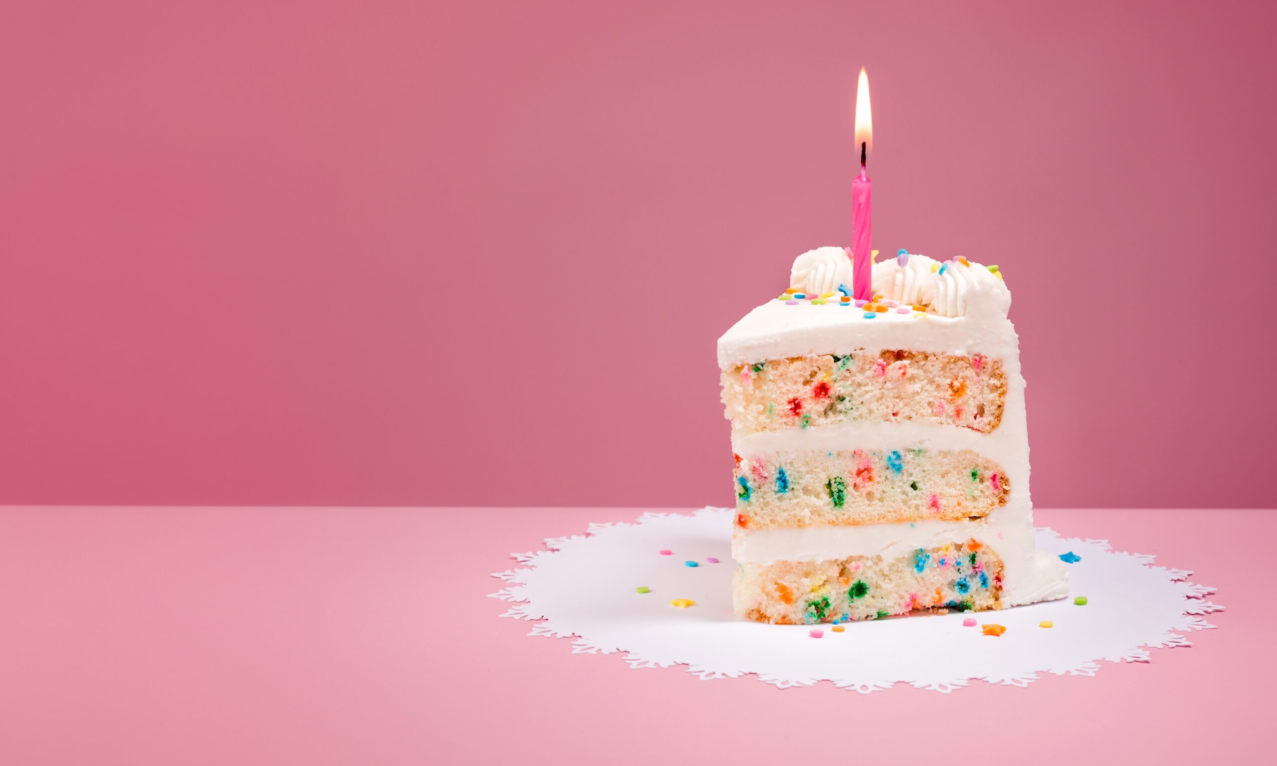 A slice of funfetti cake with vanilla frosting with one birthday candle sitting against a pink backdrop.