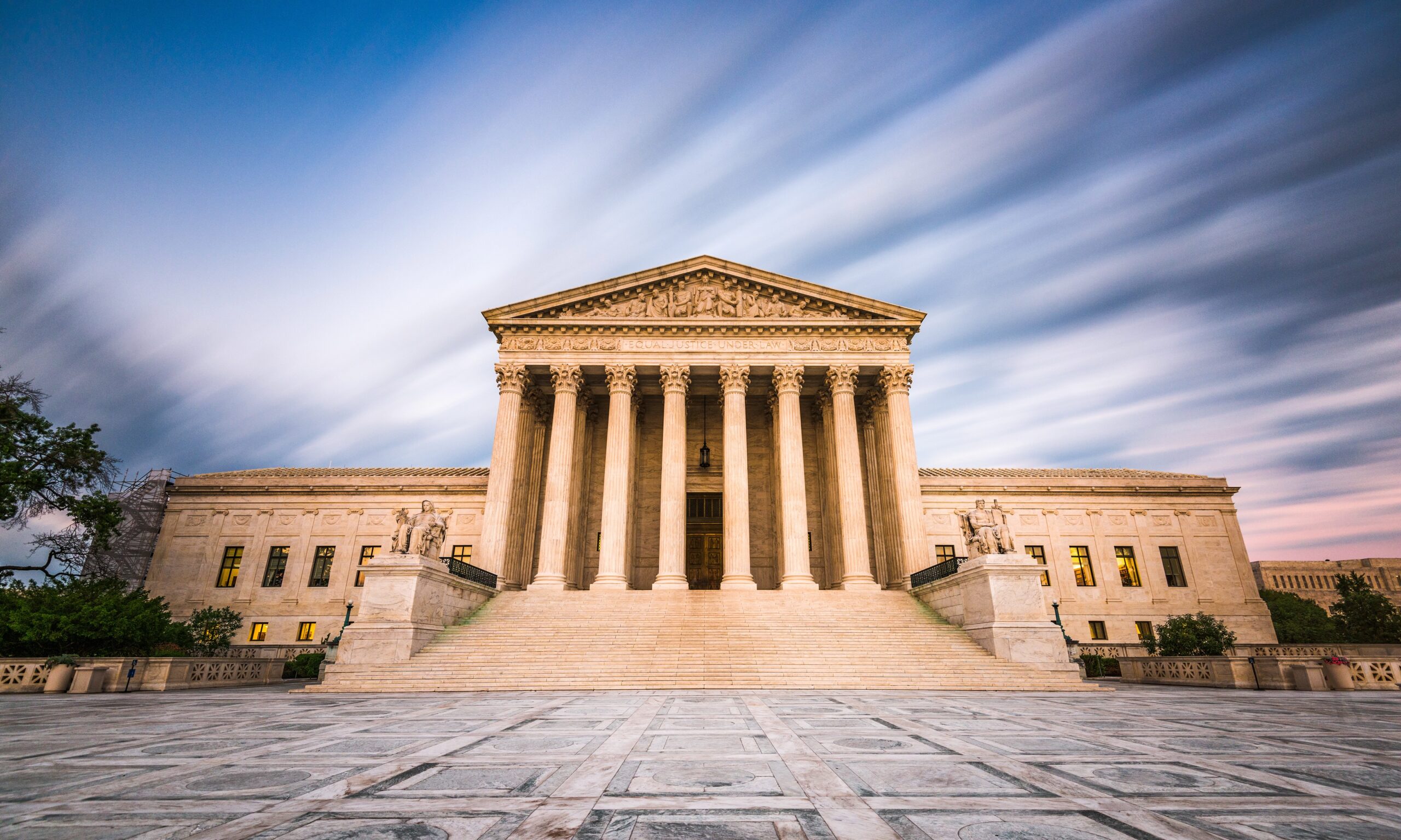 A photograph of the United States Supreme Court Building in Washington DC, USA. The building is white and has columns in the front. The sky is striped with clouds.