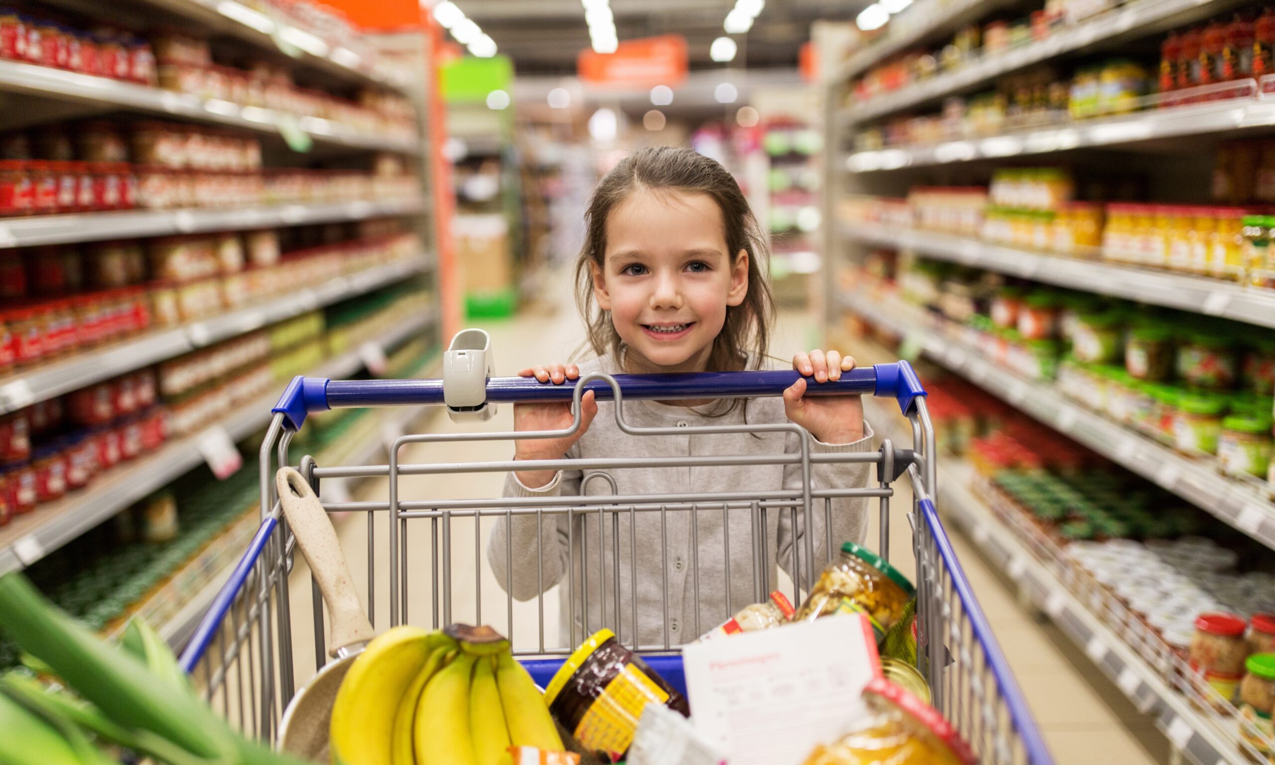 A young girl pushes a shopping cart full of groceries, including bananas and some jars, in a grocery store aisle that's slightly blurred behind her.