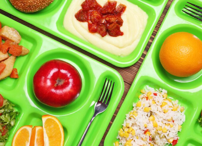 A photograph of multiple bright green cafeteria trays set close together featuring apples, oranges, grain dishes, rice and salads. Brightly colored image.