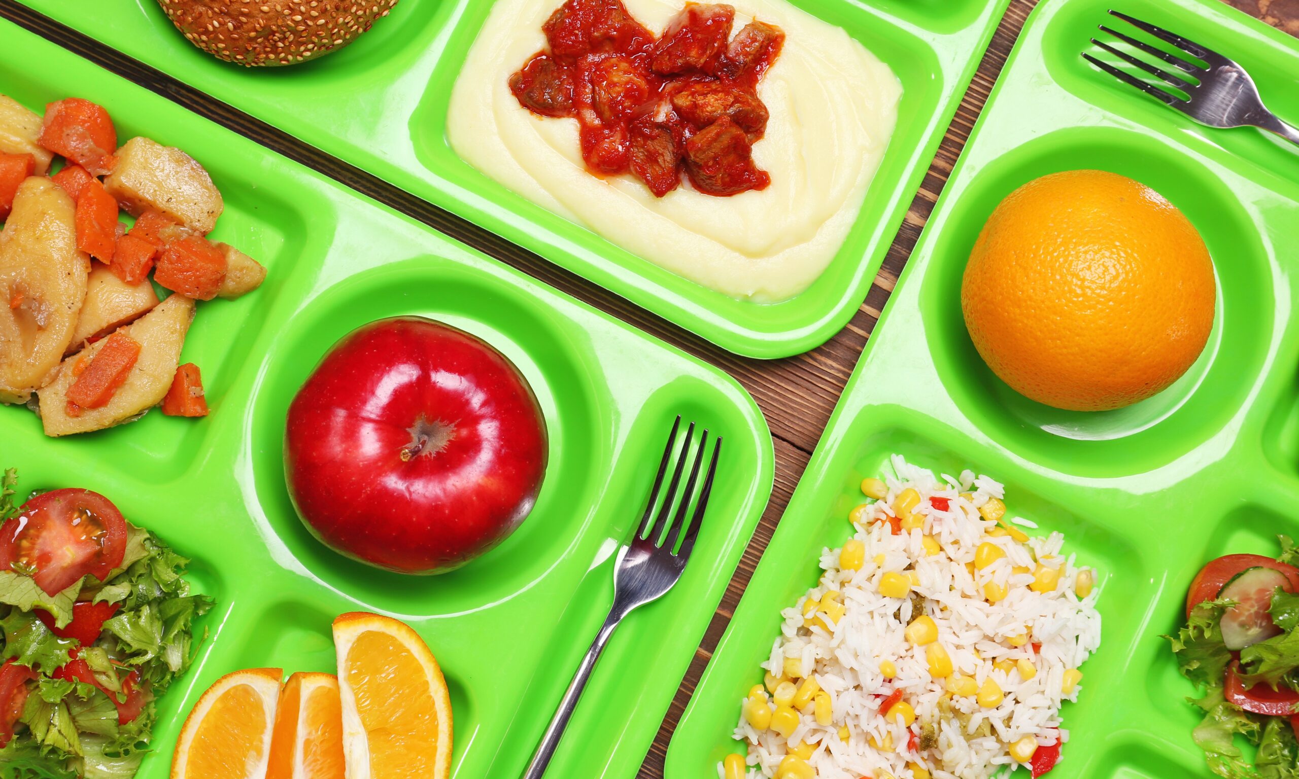 A photograph of multiple bright green cafeteria trays set close together featuring apples, oranges, grain dishes, rice and salads. Brightly colored image.