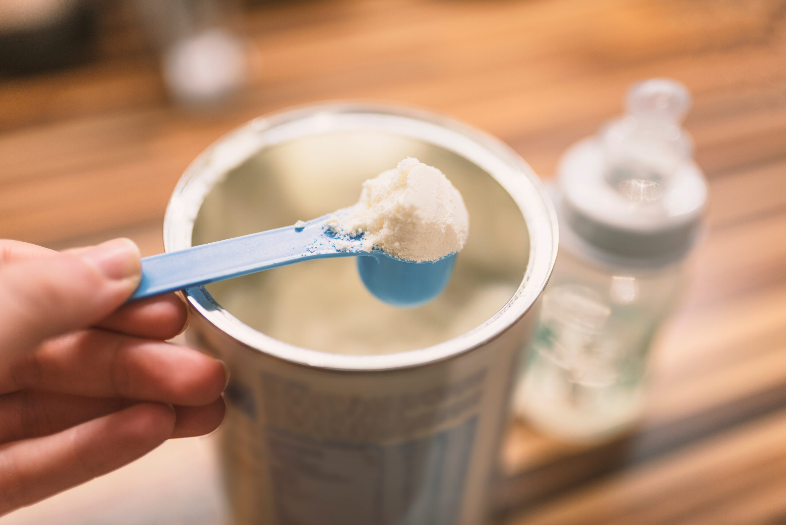 A photo of an open infant formula can with a hand holding a powdered formula scoop on top of the can.