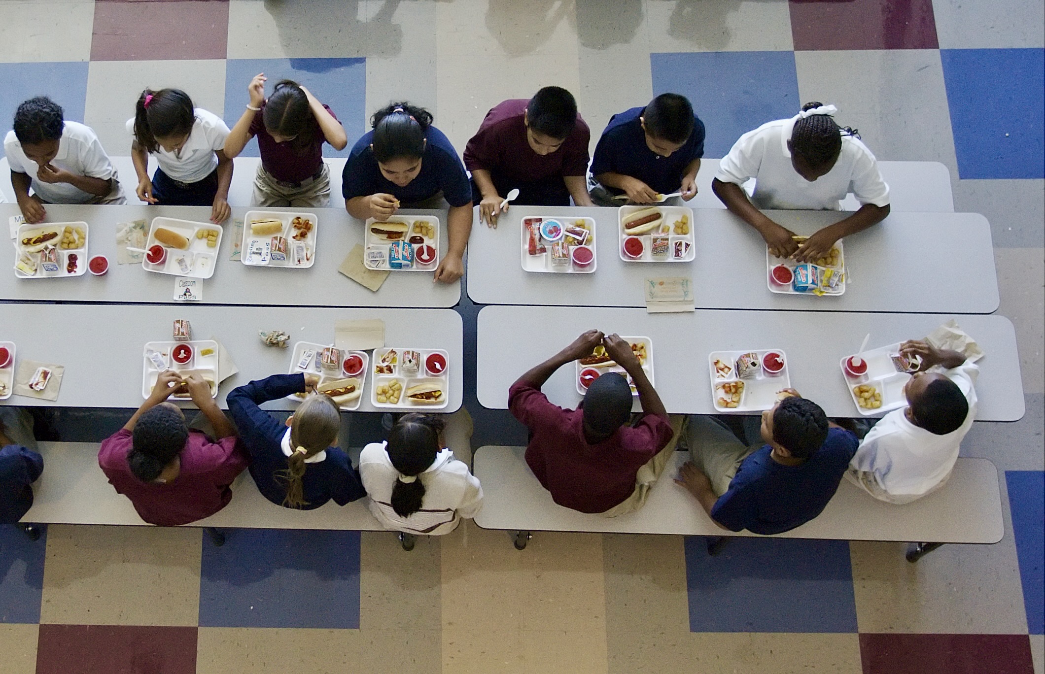 A ceiling shot looking down on a cafeteria table with children eating lunch off of cafeteria trays.