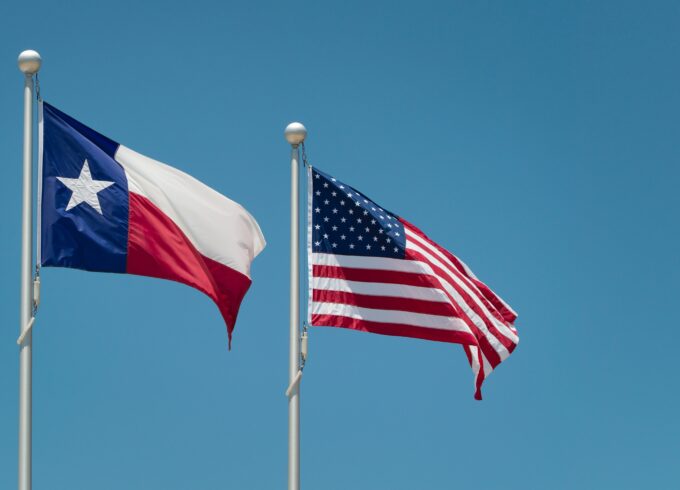 The Texas state flag flying next to the American flag against a blue sky.