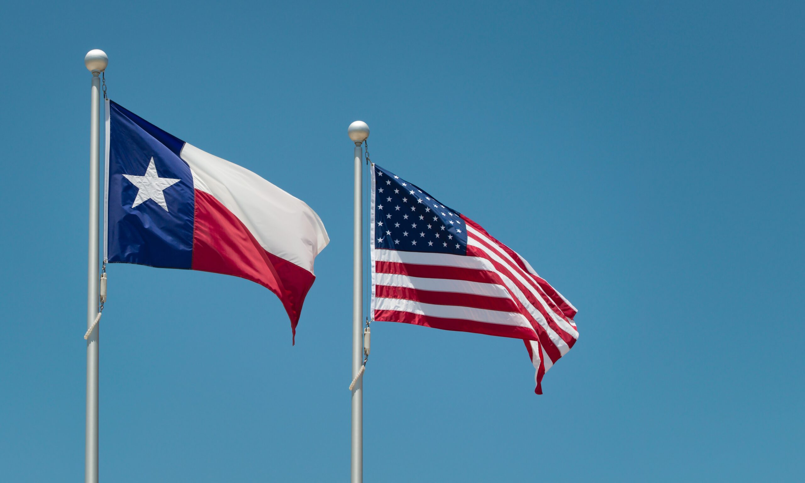 The Texas state flag flying next to the American flag against a blue sky.