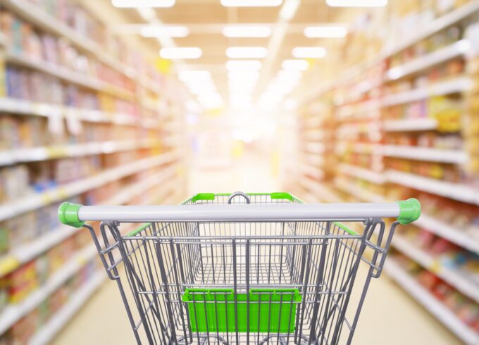 Photo of supermarket aisle product shelves interior blur background with empty shopping cart in the center.