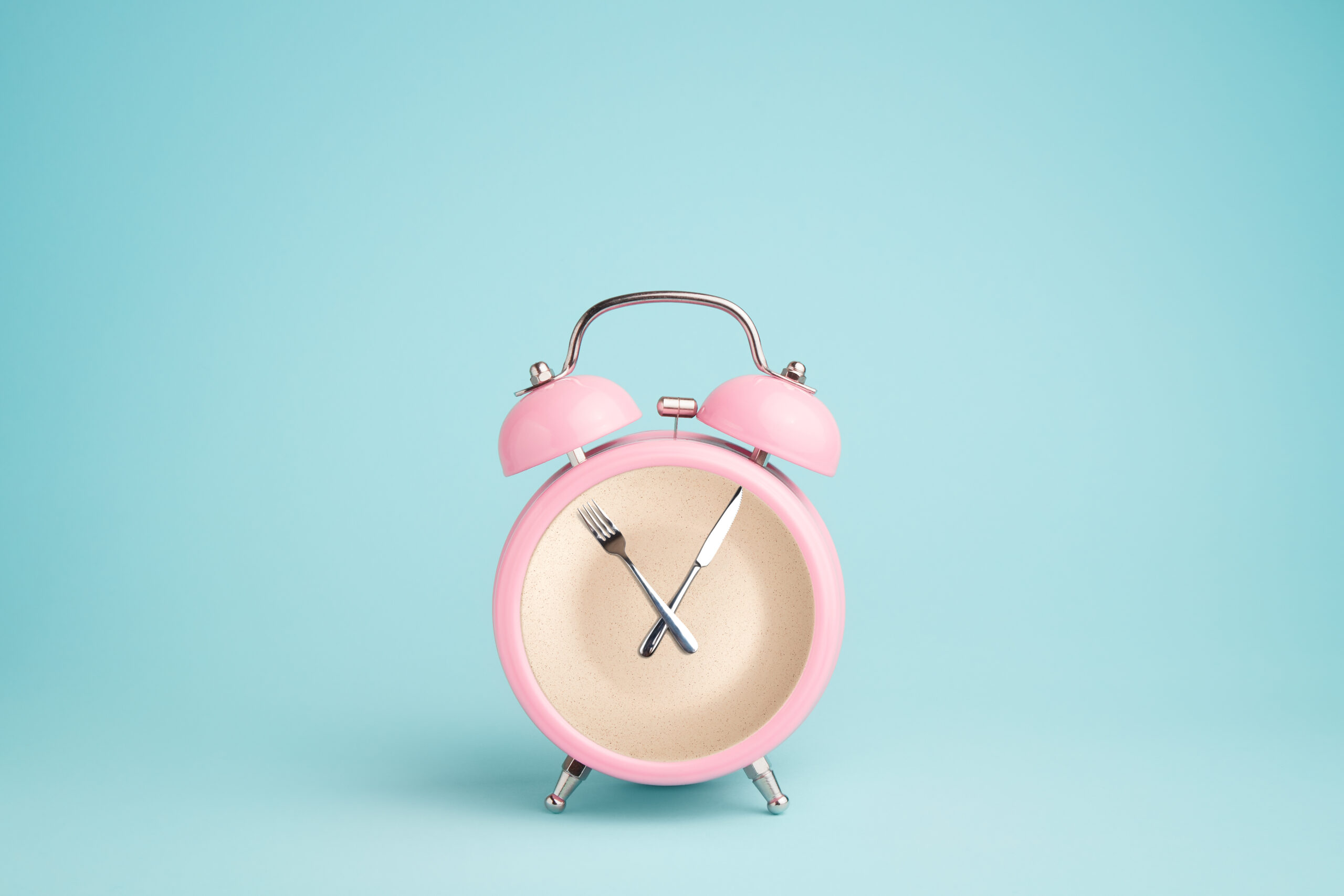 A pink alarm clock photographed against a light teal background. The clock hands are a fork and knife.