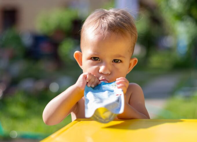 A toddler with a serious facial expression sits at a yellow table sucking on a silver snack pouch. The background is blurred but it appears to be greenery.