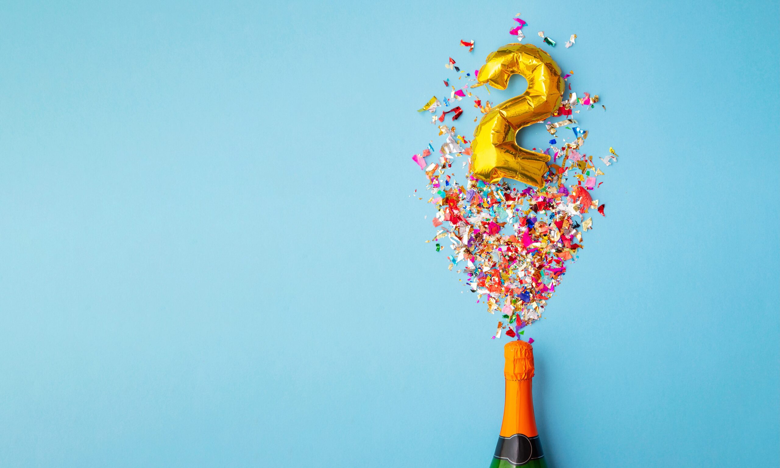 A light blue background behind a champagne bottle with confetti and a gold number two balloon made to look like it's coming out of the bottle.