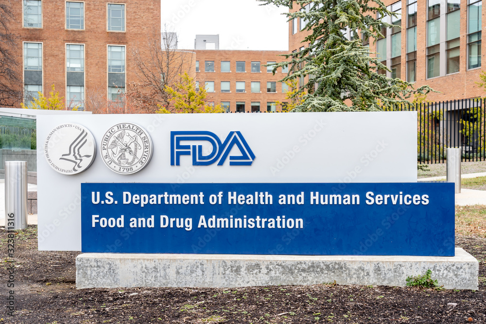 A large concrete sign reading FDA in large blue letters and below it U.S. Department of Health and Human Services Food and Drug Administration. Seals of HHS and Public Health Service in he upper left hand corner with brick buildings and trees in the background.