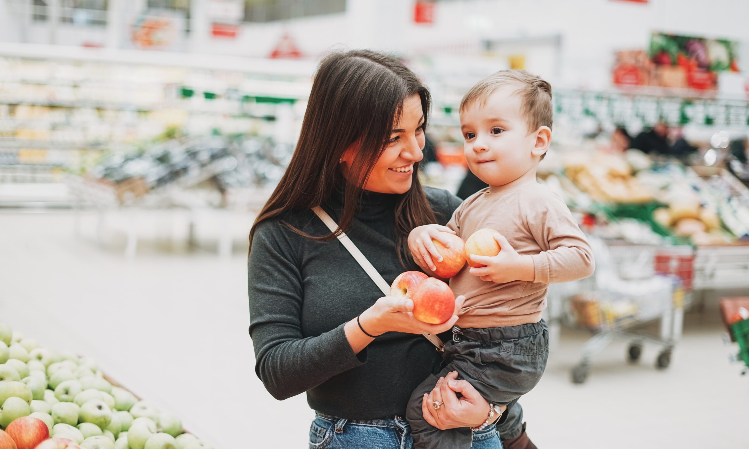 A mom with long brown hair holds a toddler in the produce section of a grocery store. They are both holding peaches.