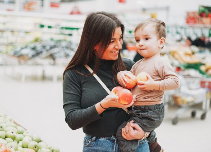 A woman wearing a turtle neck is holding a toddler in the produce section of the grocery store. They are both holding apples.