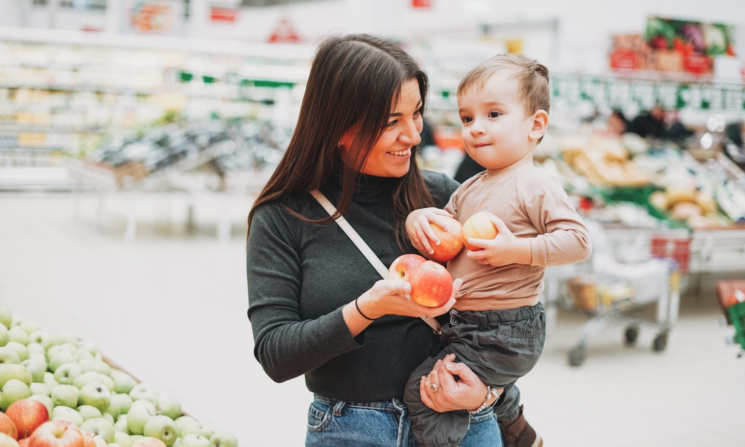 A woman wearing a turtle neck is holding a toddler in the produce section of the grocery store. They are both holding apples.