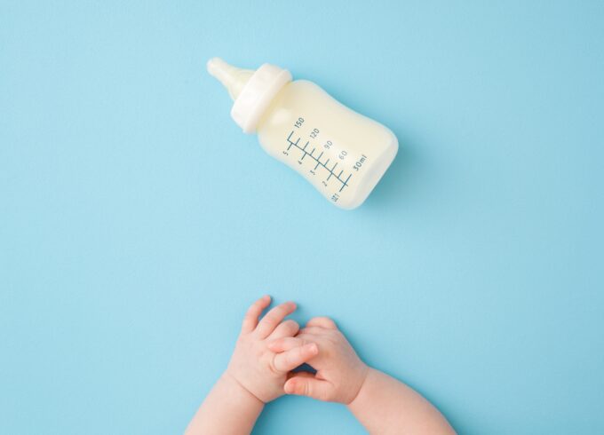 Infant hands and bottle of milk on light blue table background. Feeding time. Pastel color. Closeup. Point of view shot. Top down view.