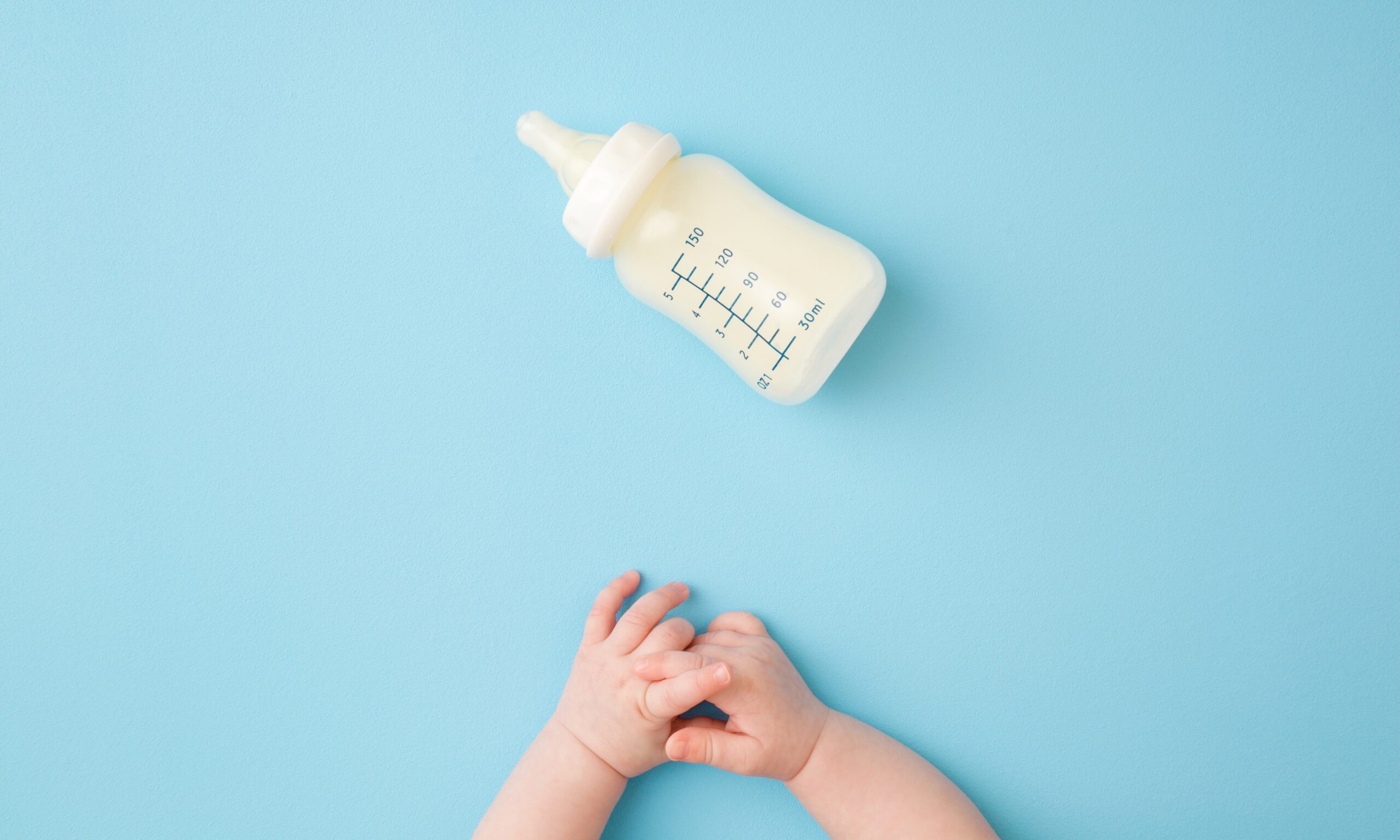 Infant hands and bottle of milk on light blue table background. Feeding time. Pastel color. Closeup. Point of view shot. Top down view.