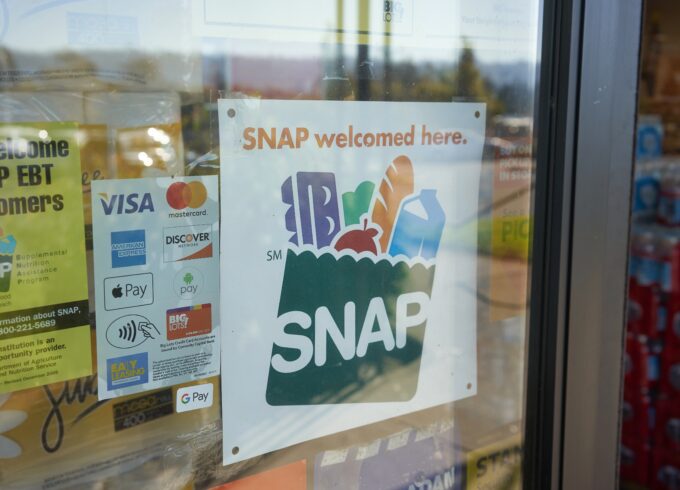 A window features a sign that reads SNAP accepted here. The sign includes a logo that's a grocery bag that reads "SNAP" on it filled with various grocery items.