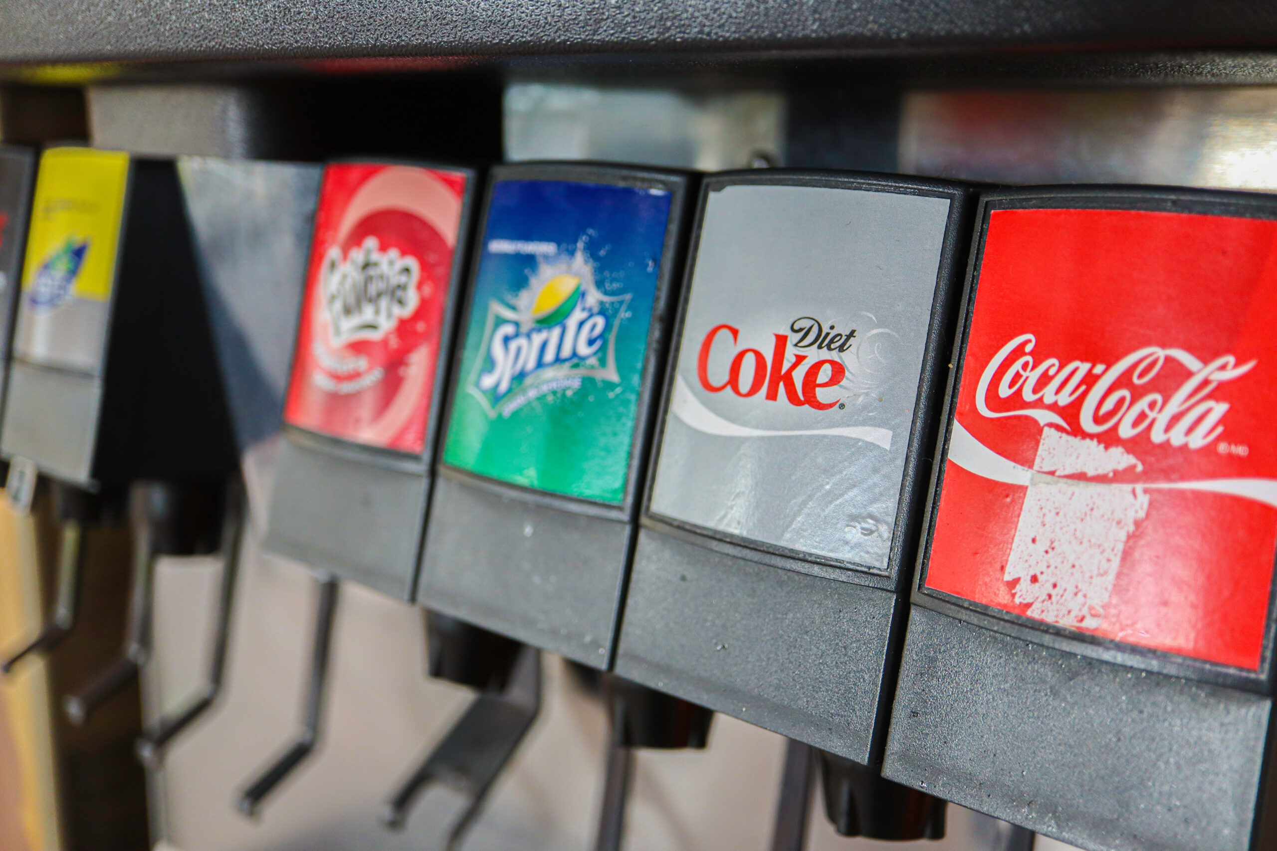 A photograph of a soda fountain focused on the brand names: Coca-Cola, Diet Coke, etc.