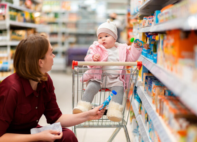 A woman crouches next to a shopping cart in a grocery aisle. A baby is sitting in the grocery cart reaching for baby food.