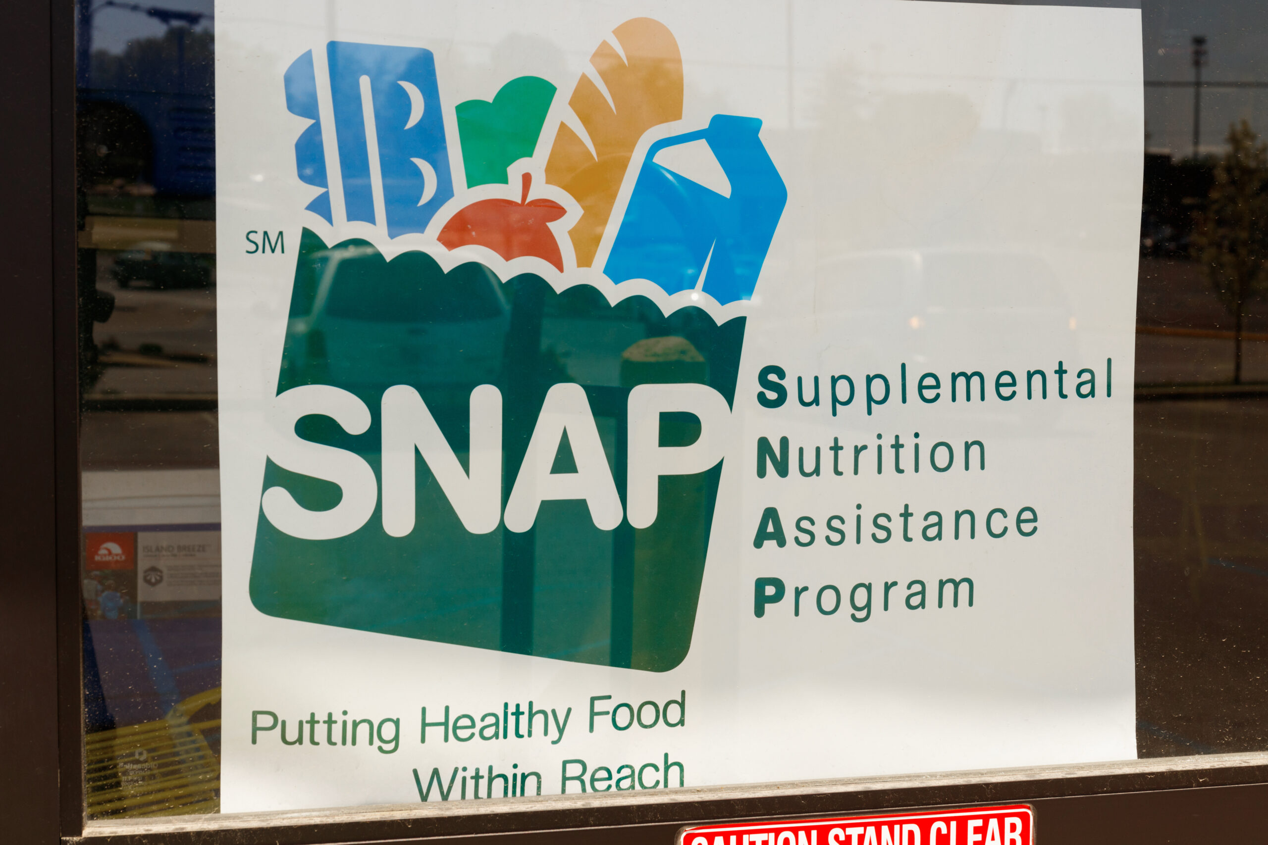 A window sign features the SNAP logo, which is a green grocery bag filled with eggs, produce, bread and milk. It says Supplemental Nutrition Assistance Program spelled out next to the logo.