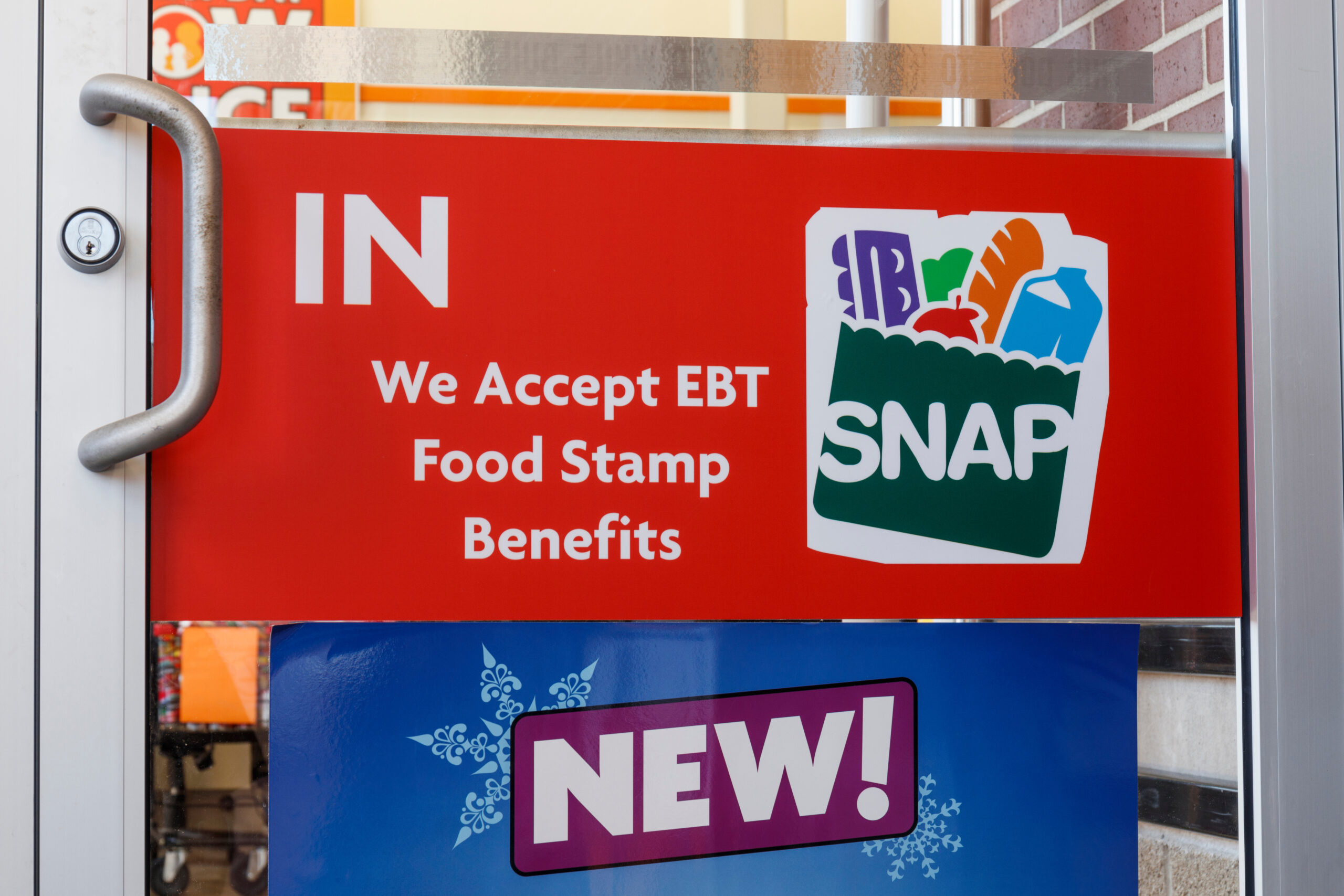 A red sign on a retailers glass door says: IN. We Accept EBT Food Stamp Benefits