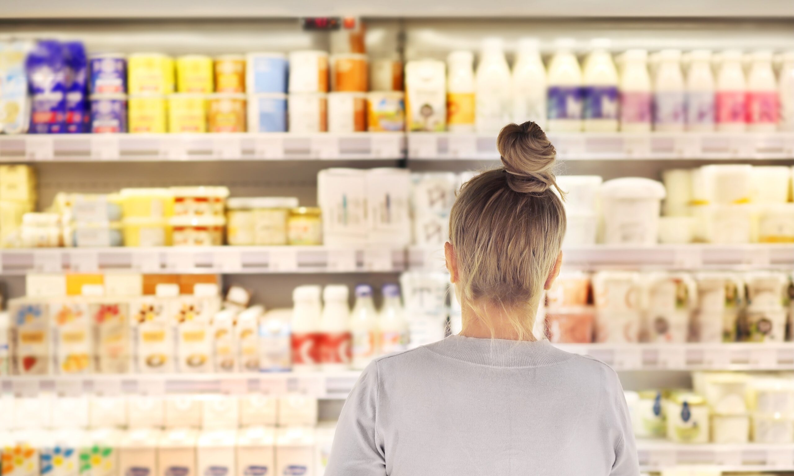 A woman with blonde hair stands with her back to the camera - her hair is in a bun and she's facing a dairy aisle that's out of focus.