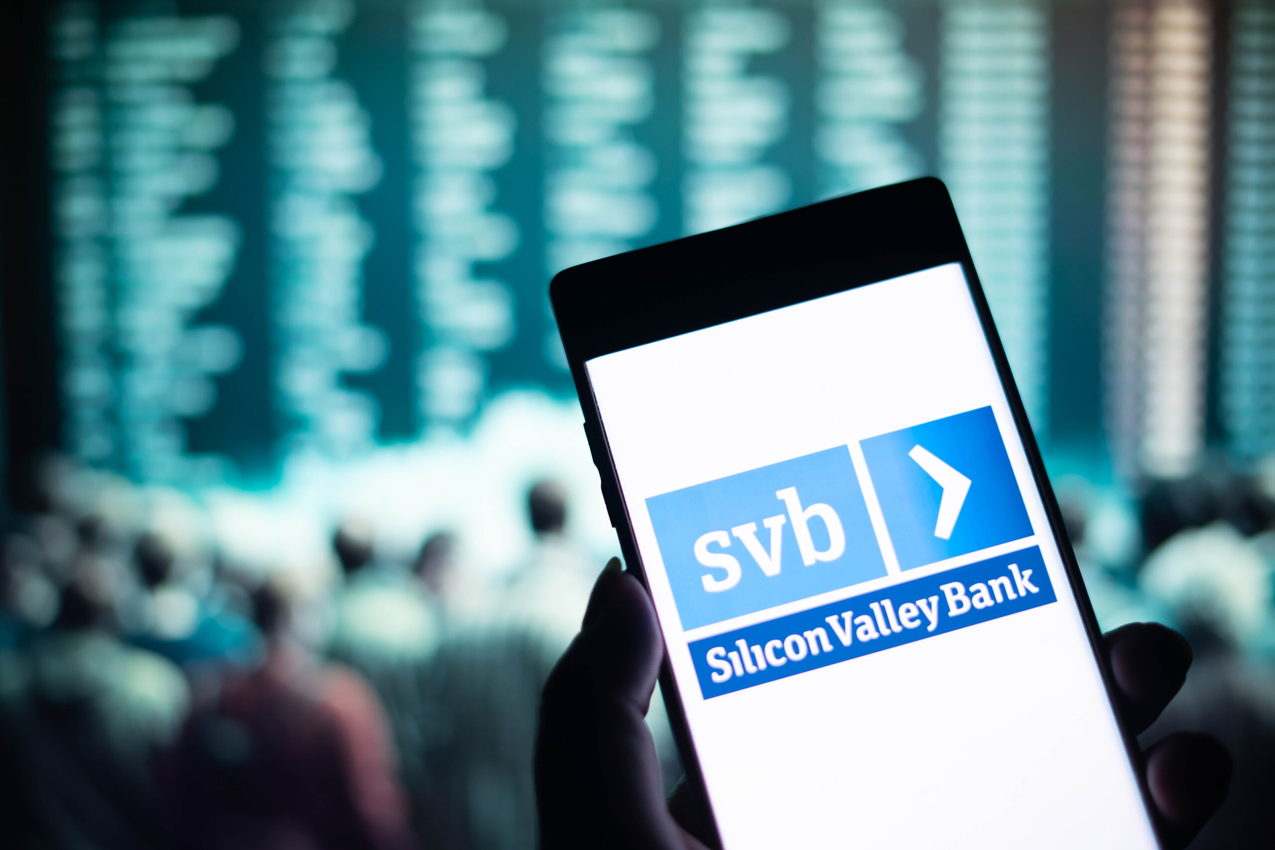 The Silicon Valley Bank logo appears on a smart phone held in a hand -- in the background is blurred out financial charts.