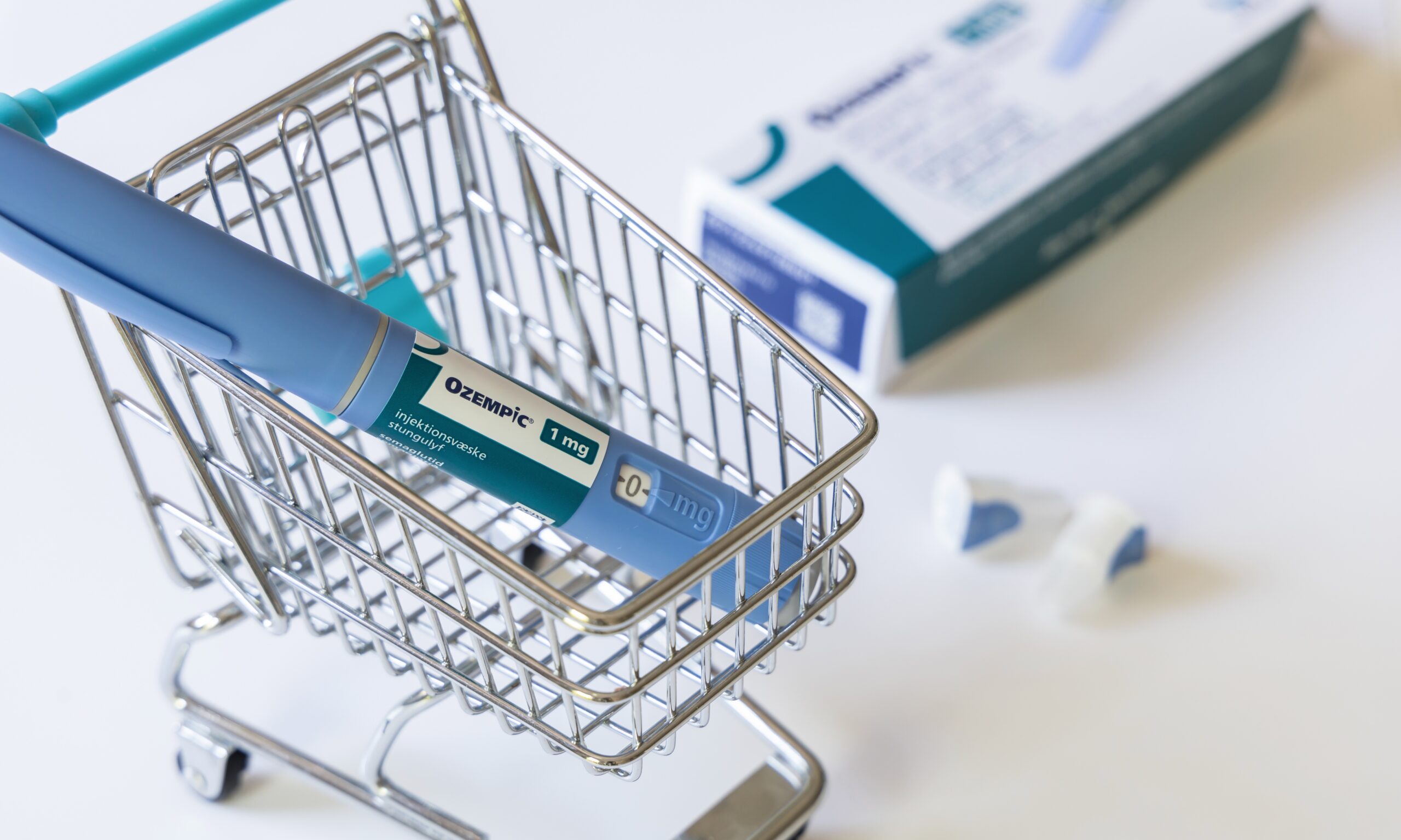An Ozempic injectable pen is shown inside a small metal toy grocery cart. There's a drug box sitting out of focus behind the cart.