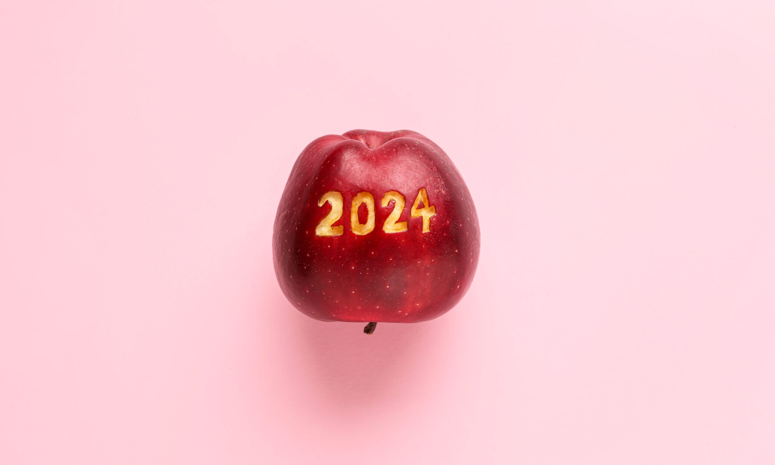 A red apple sits upside down against a light pink background. 2024 is carved into the apple.