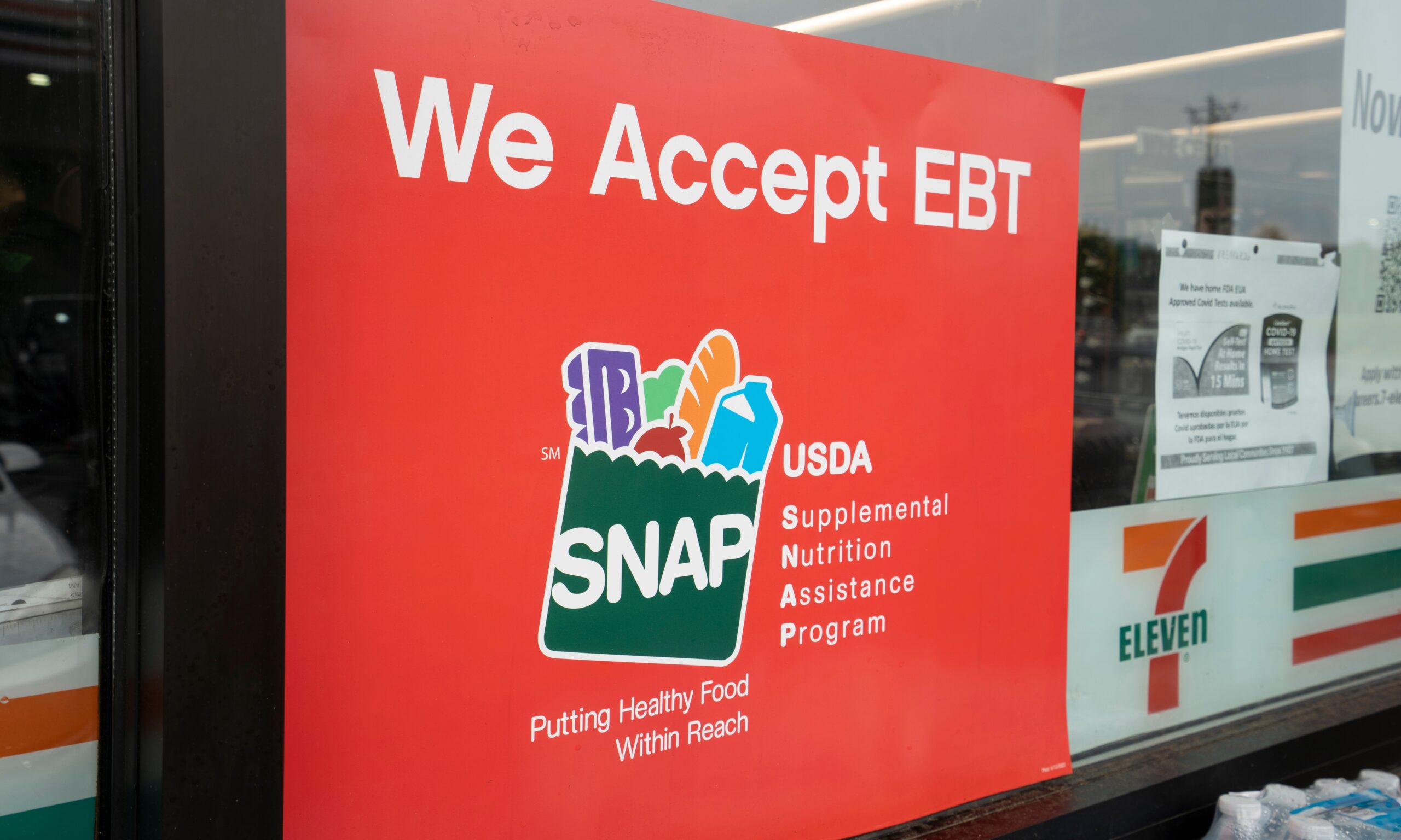 A photograph of a red sign on a 7 Eleven door that says "We Accept EBT" with the USDA SNAP logo on it.