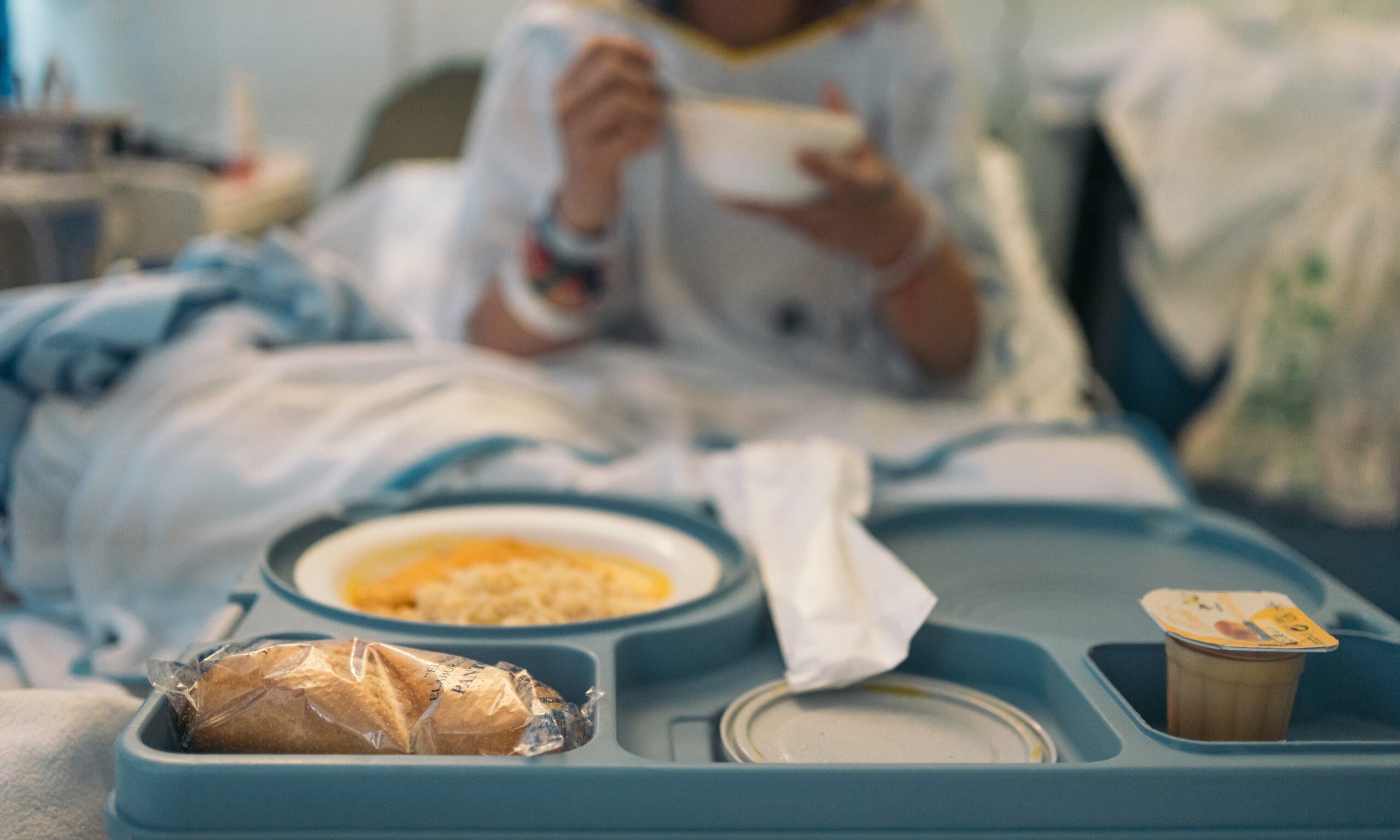 Close-up of untouched hospital food tray with an unidentifiable woman in the background eating soup.