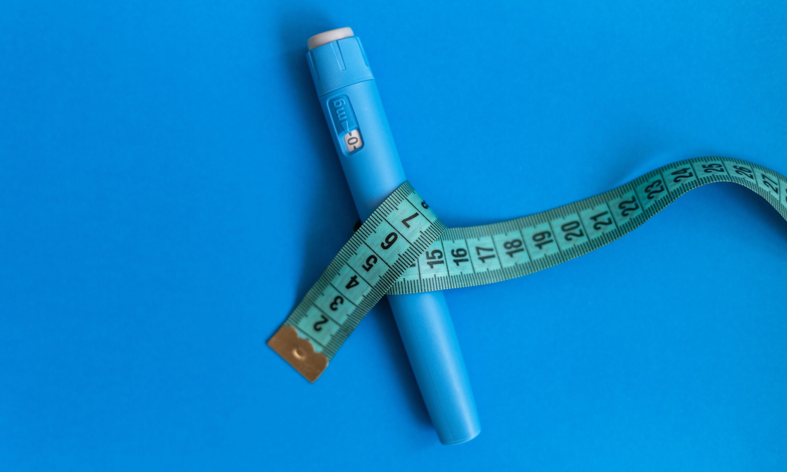 A blue Ozempic pen sits on top of a blue background. Wrapped around the pen is a green measuring tape.