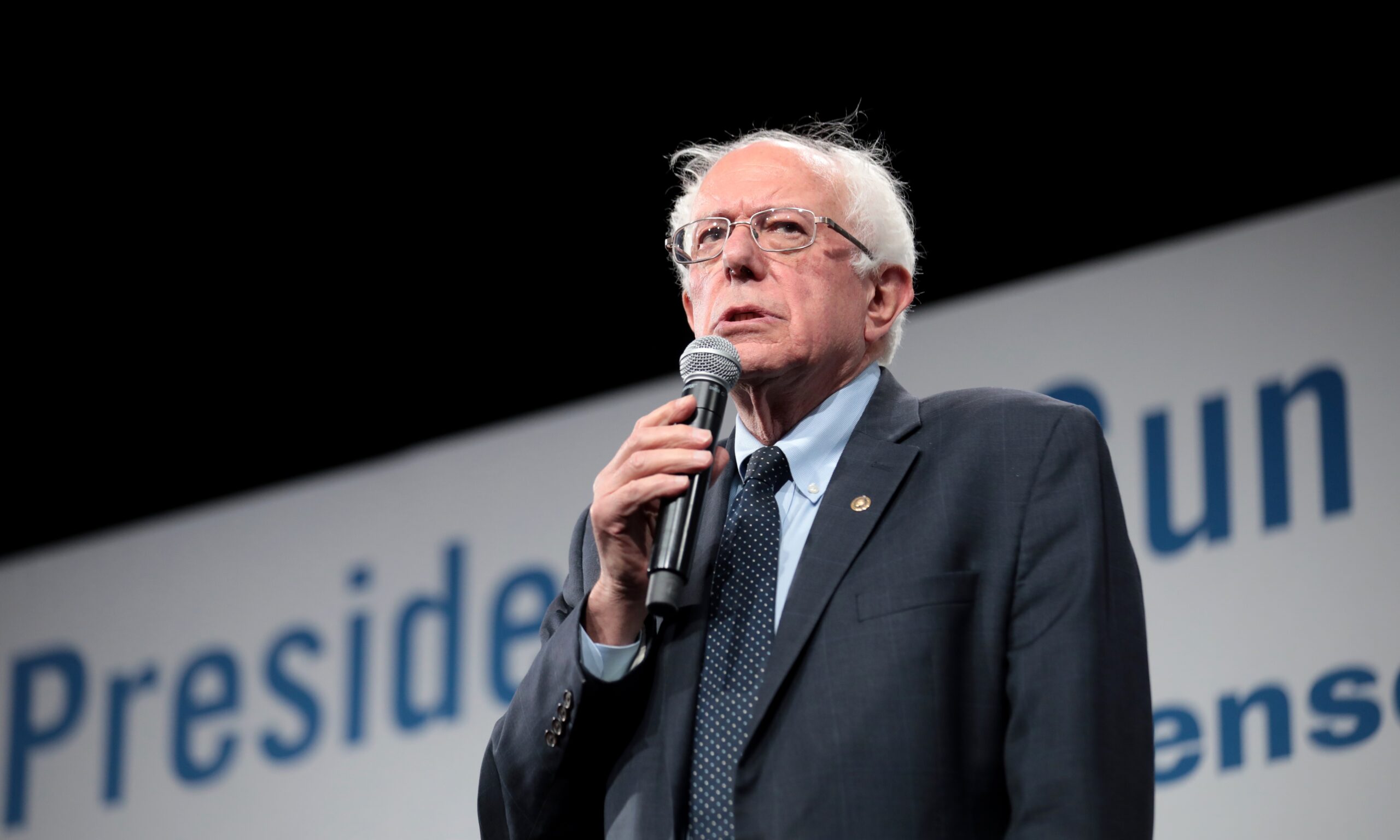 Bernie Sanders is pictured holding a microphone, wearing a dark suit and tie - making a speech in front of a dark background.