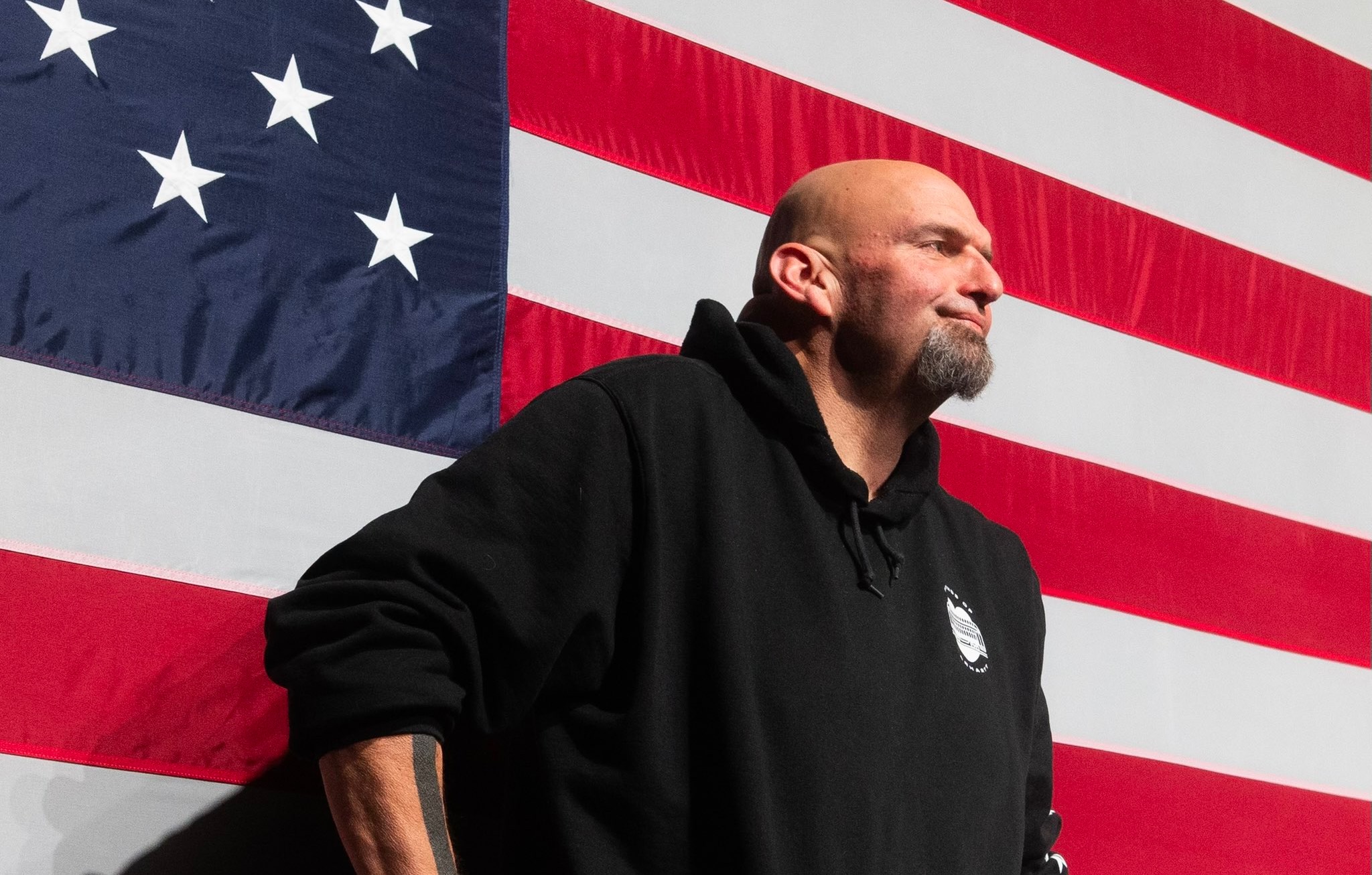 A photograph of Senate candidate John Fetterman standing in front of an American flag wearing a black hoodie.