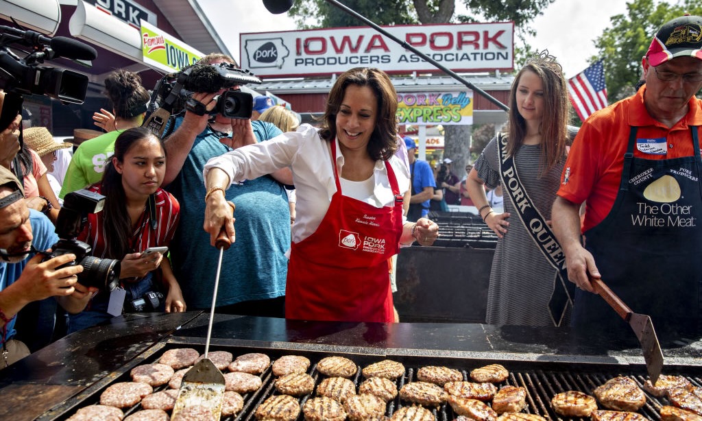 A photograph of Kamala Harris flipping pork on a large grill surrounded by reporters at the Iowa State Fair in 2019. She's wearing a red branded apron and a white button up shirt.