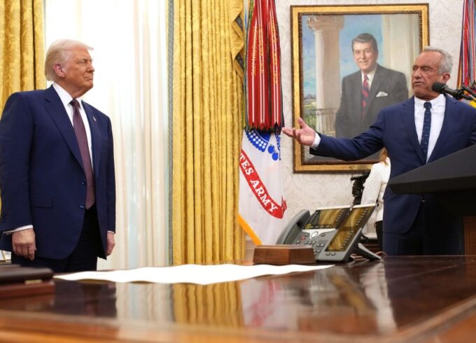 Robert F. Kennedy Jr. speaks alongside President Donald Trump in the Oval Office. The desk is in the foreground and both speakers are standing in the back wearing dark suits.