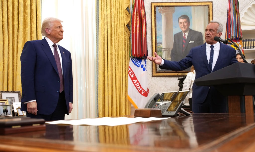 Robert F. Kennedy Jr. speaks alongside President Donald Trump in the Oval Office. The desk is in the foreground and both speakers are standing in the back wearing dark suits.