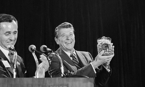 A black and white photograph of Ronald Reagan standing at a podium holding a crystal jar of jelly beans.