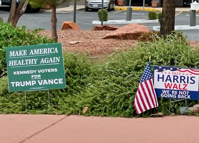 A green "Make America Healthy Again" Kennedy Voters for Trump Vance sign competes with a Harris Walz "We're not going back" sign.